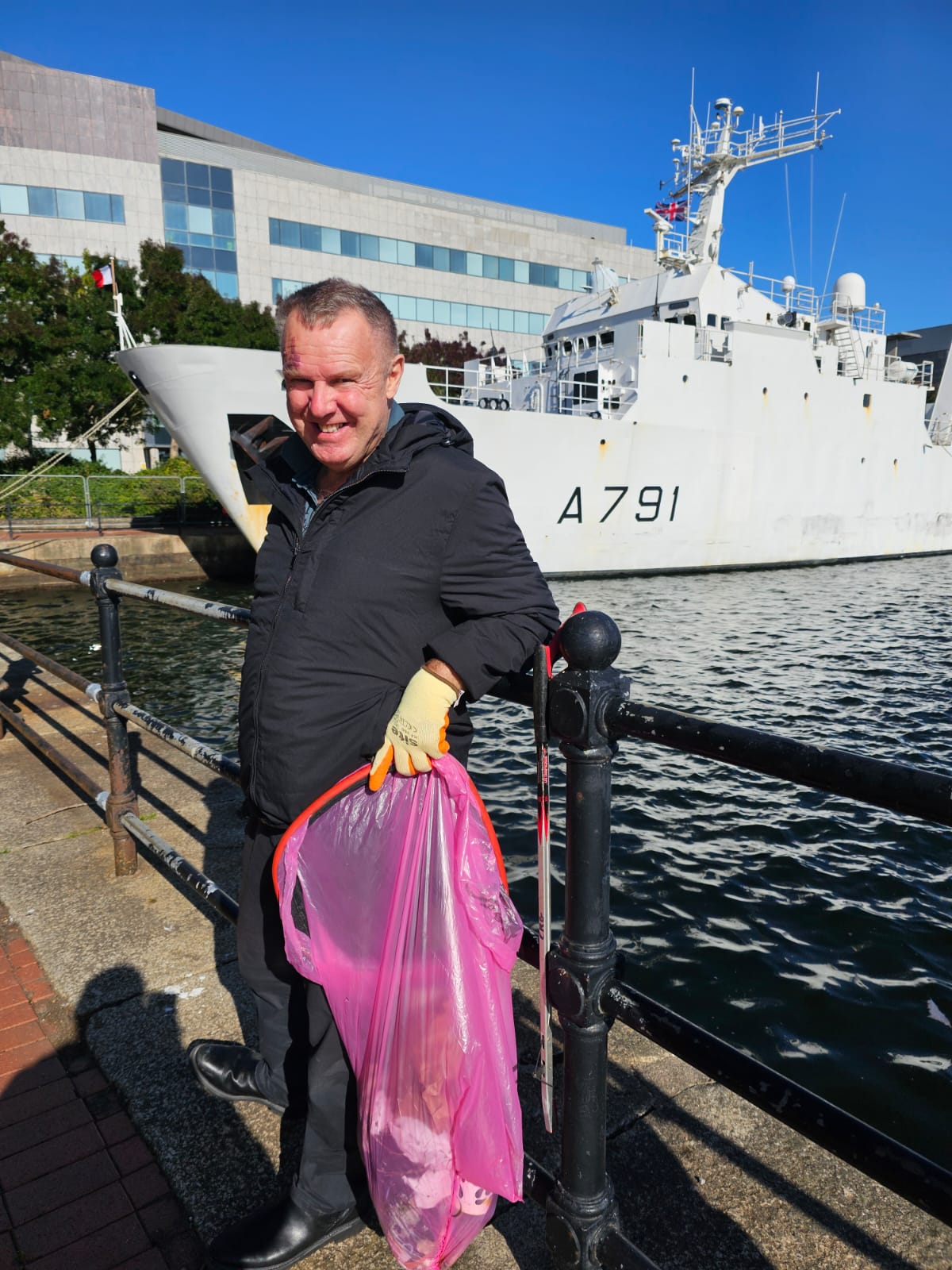 Rob on his second bag at the Cardiff Bay litter pick. Rob is supported by Shared Lives. He's smiling at the camera with a war ship behind him.