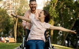 Woman in a wheelchair being pushed through a park by a man. Both look very happy and the woman has her arms outstretched to the side.d 