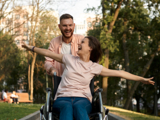 Woman in a wheelchair being pushed through a park by a man. Both look very happy and the woman has her arms outstretched to the side.d 