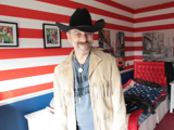 Trevor stood in his bedroom with a cowboy hat on. The wall paper behind him is red and white horizontal stripes.