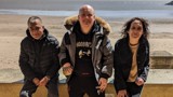 Three smiling residents of Ty Cornel, two men and a woman, sitting on a wall with a view of the beach and the ocean behind them. 