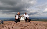 A man and a woman stood next to the Pen Y Fan sign on top of the mountain. 
